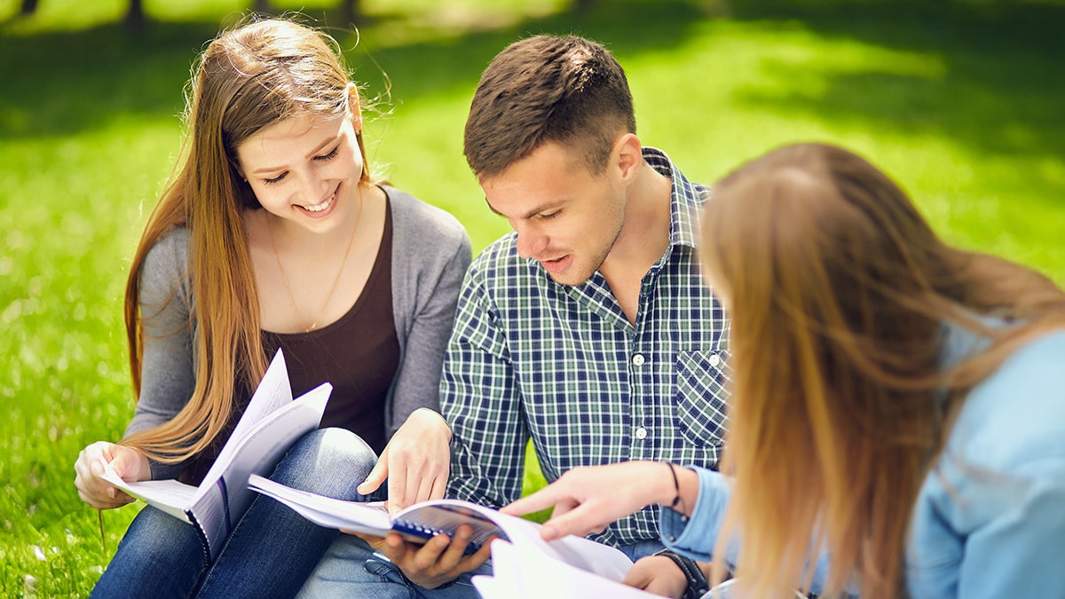Three university students sitting on grass and reading course notes together.