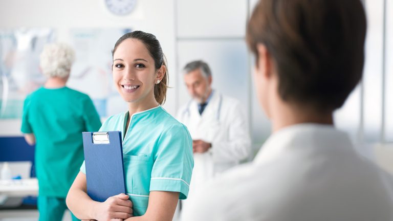 Smiling registered nurse holding a clipboard in a hospital clinic with healthcare staff nearby.