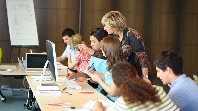 Secondary school teacher helping students work at desktop computers during a classroom learning session.