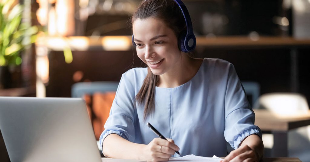 Smiling young woman with headphones taking notes in front of laptop