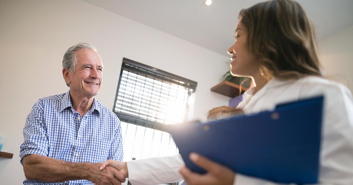 Woman wearing white coat and holding clipboard shaking hands with an old man patient or client