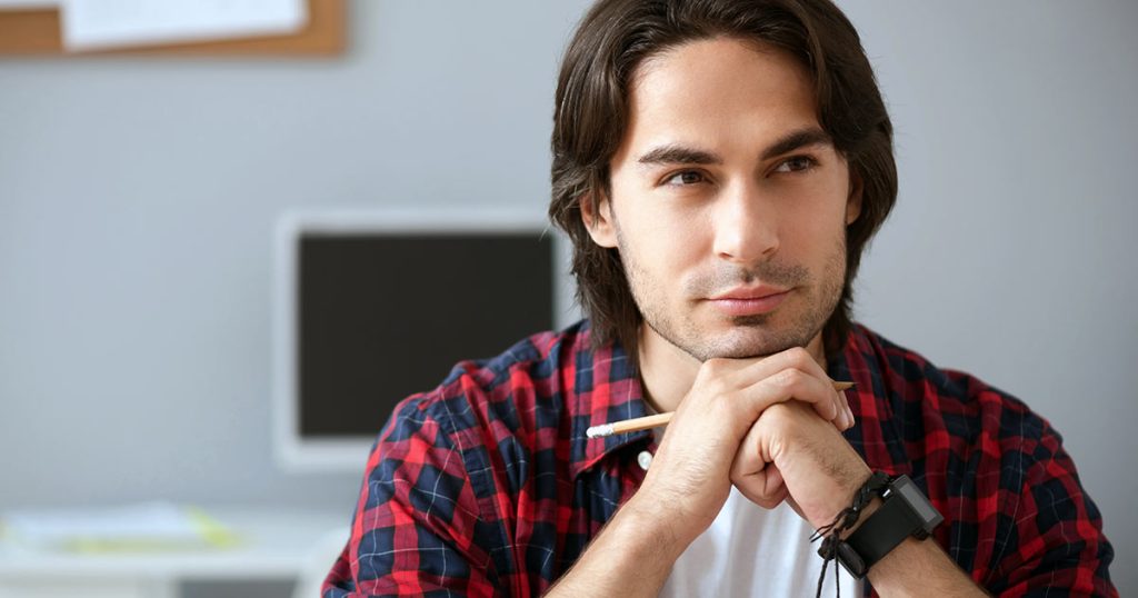 Young man in check shirt in contemplative pose holding pen in front of computer