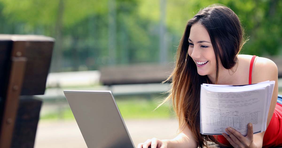 Woman on outdoor table lying on stomach studying with laptop and notebook