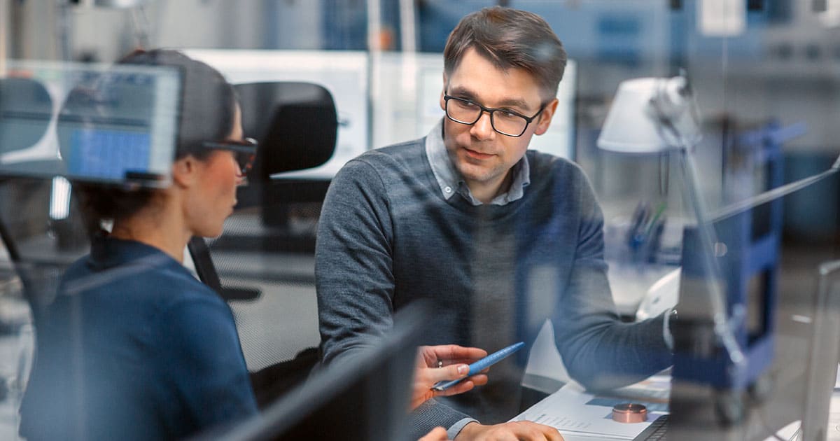 Two work colleagues with glasses at a workstation talking