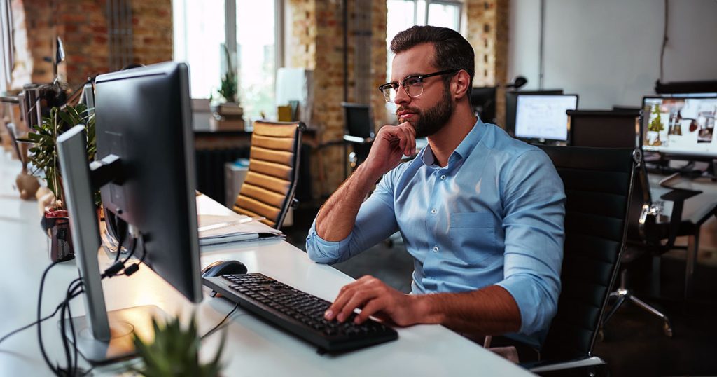 Young bearded man working on computer touching chin sitting in office
