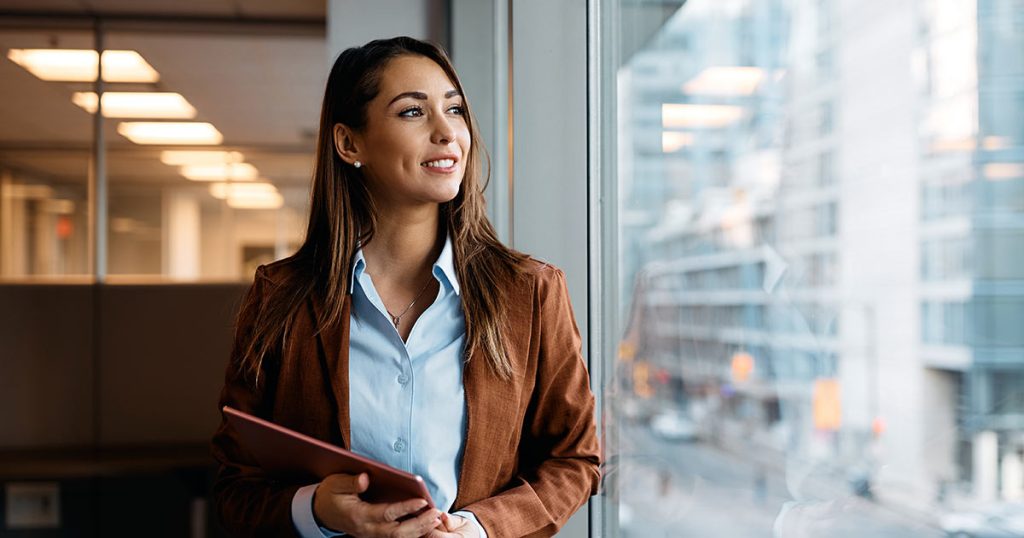 Young professional woman looking outside office building window while holding touchpad