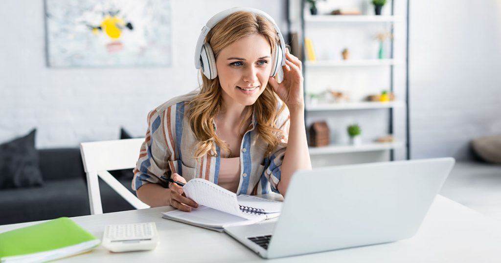 Young woman taking notes from laptop wearing headphones