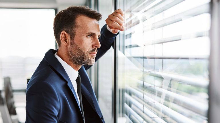 Business minded executive looking thoughtfully through office blinds while reflecting on strategy, opportunity, and decision-making.