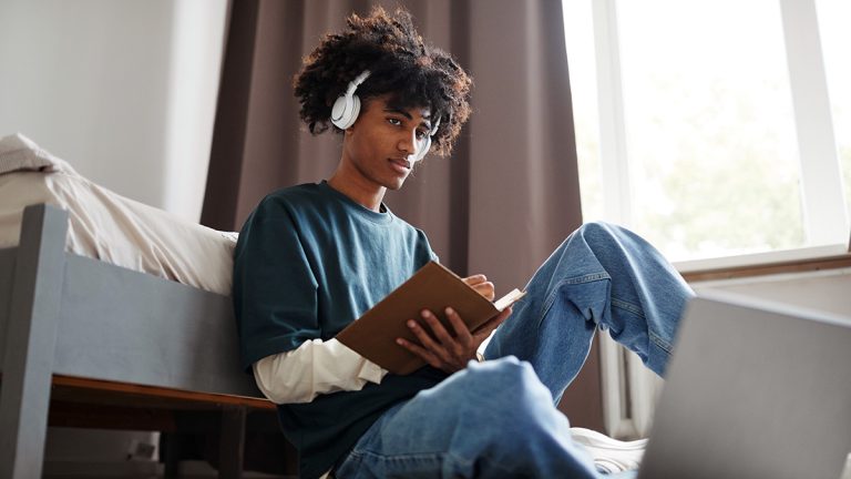 College student wearing headphones and writing in a notebook beside a laptop in a dorm room