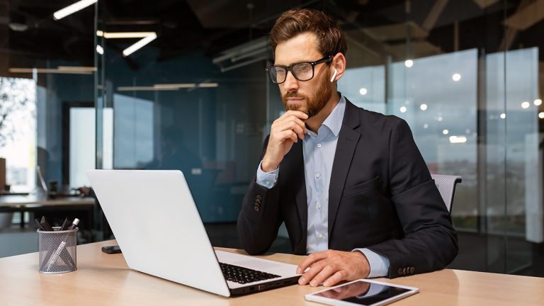 Data analyst wearing glasses and earbuds concentrating on a laptop at an office desk.