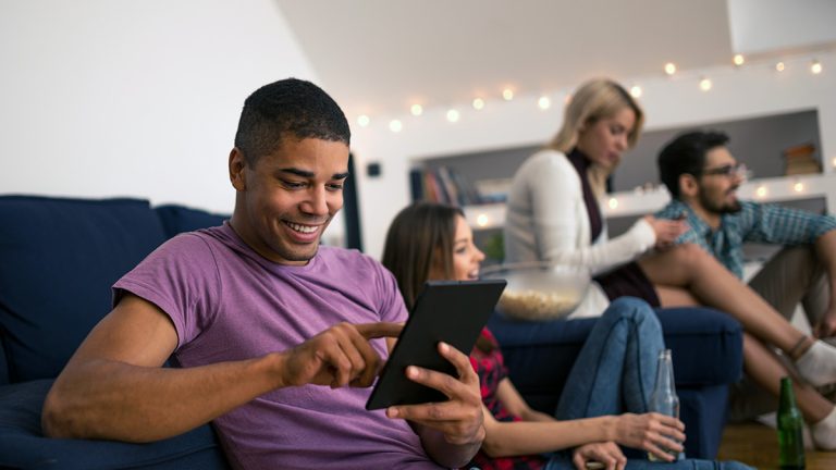 Young man using a tablet on a sofa while friends socialise nearby.