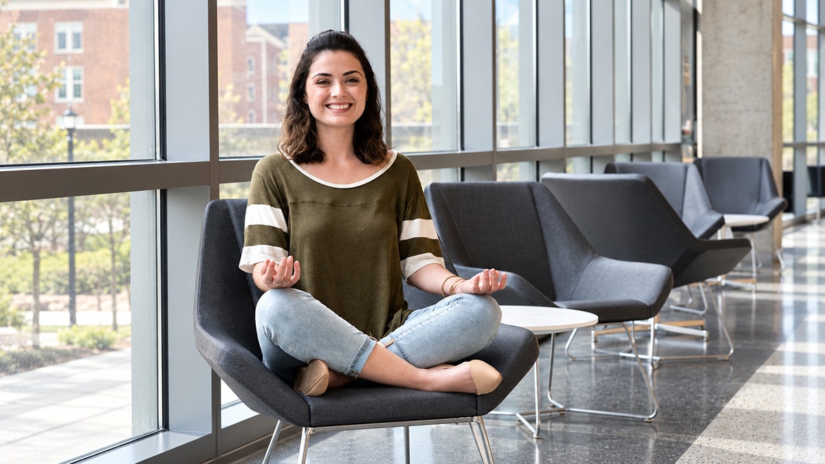 College student sitting cross-legged on a lounge chair, smiling with her palms facing upward.