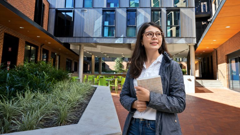 International student holding a notebook in a modern Sydney accommodation courtyard.