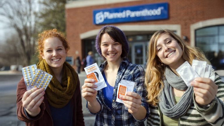 Three female college students holding different birth control options and smiling outdoors near a health clinic