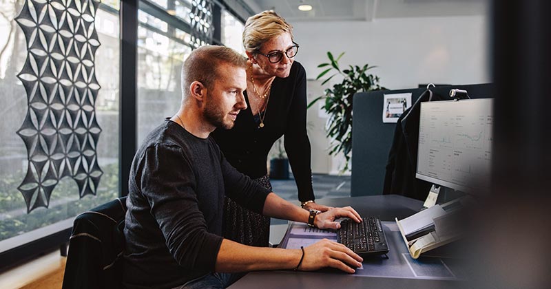 Female professional overlooking work being produce by male colleague on computer